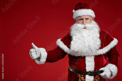 Portrait of Santa Claus in beautiful costume standing and showing thumb up isolated on red background © AnnaStills