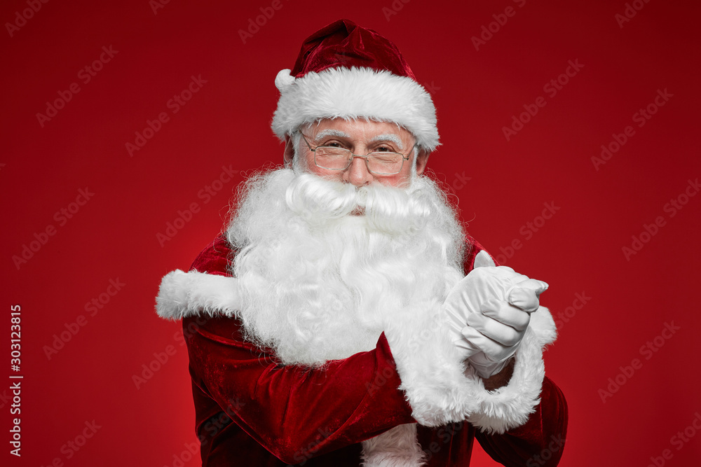Portrait of Santa Claus in eyeglasses and with white beard showing respect and looking at camera isolated on red background Portrait of Santa Claus in eyeglasses and with white beard showing respect and looking at camera isolated on red background
