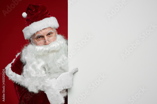 Portrait of Santa Claus with white beard looking at camera while standing near the big blank placard and pointing at it © AnnaStills