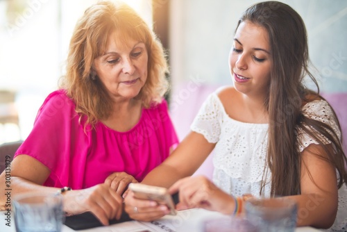 Beautiful mother and daugther sitting at restaurant using smartphone smiling