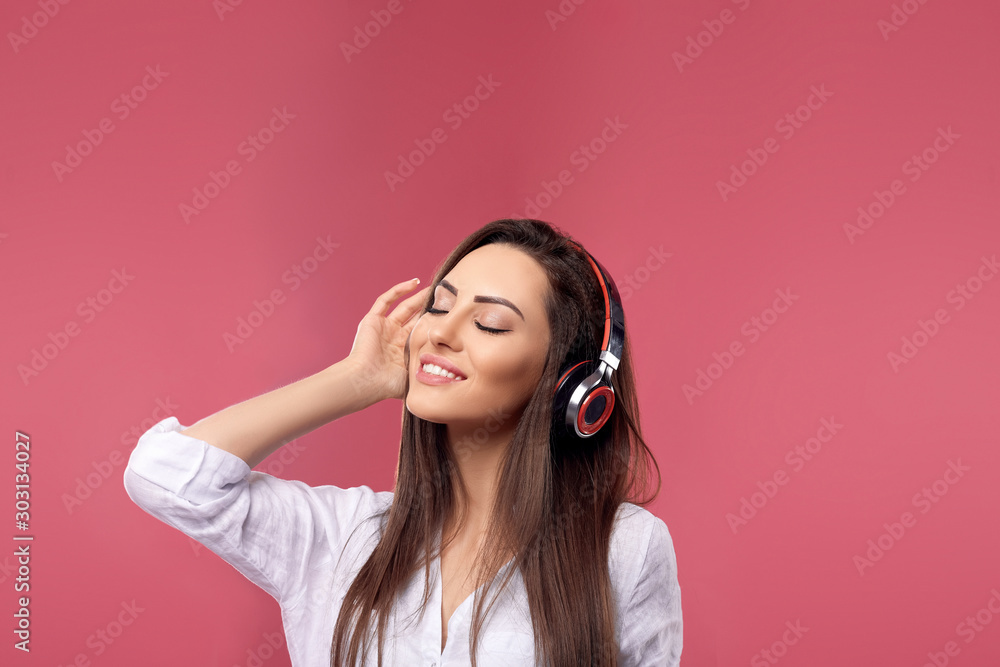 Beautiful woman smiling with smartphone in hands enjoying favorite music, via wireless headphones over pink background. Girl uses wireless earphones and dancing.. Beautiful woman smiling with smartphone in hands enjoying favorite music, via wireless headphones over pink background. Girl uses wireless earphones and dancing..