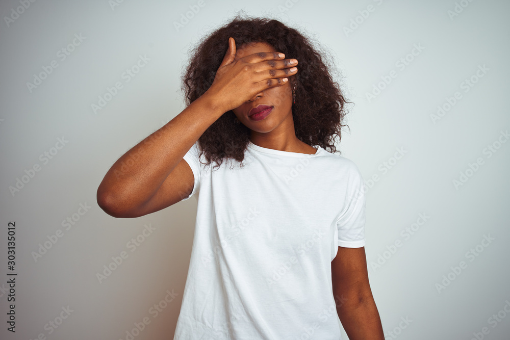 Young african american woman wearing t-shirt standing over isolated white background covering eyes with hand, looking serious and sad. Sightless, hiding and rejection concept Young african american woman wearing t-shirt standing over isolated white background covering eyes with hand, looking serious and sad. Sightless, hiding and rejection concept
