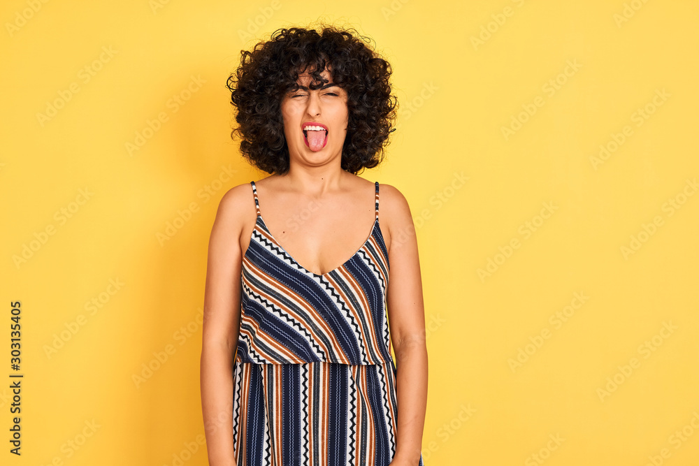 Arab woman with curly hair wearing striped colorful dress over isolated yellow background sticking tongue out happy with funny expression. Emotion concept. Arab woman with curly hair wearing striped colorful dress over isolated yellow background sticking tongue out happy with funny expression. Emotion concept.