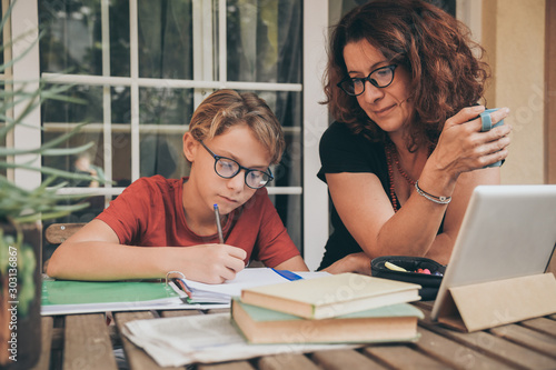 Φωτογραφία Young student doing homework at home with school books newspaper, digital pad helped by his mother