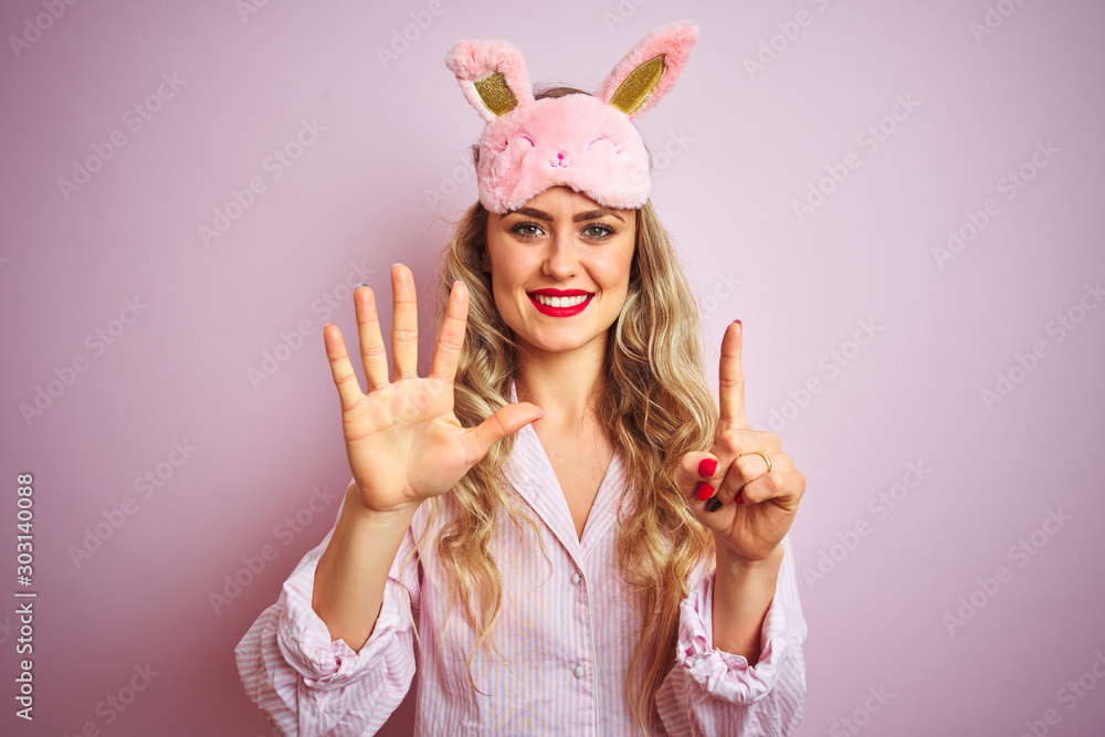 Young beautiful woman wearing pajama and sleep mask over pink isolated background showing and pointing up with fingers number six while smiling confident and happy. Young beautiful woman wearing pajama and sleep mask over pink isolated background showing and pointing up with fingers number six while smiling confident and happy.