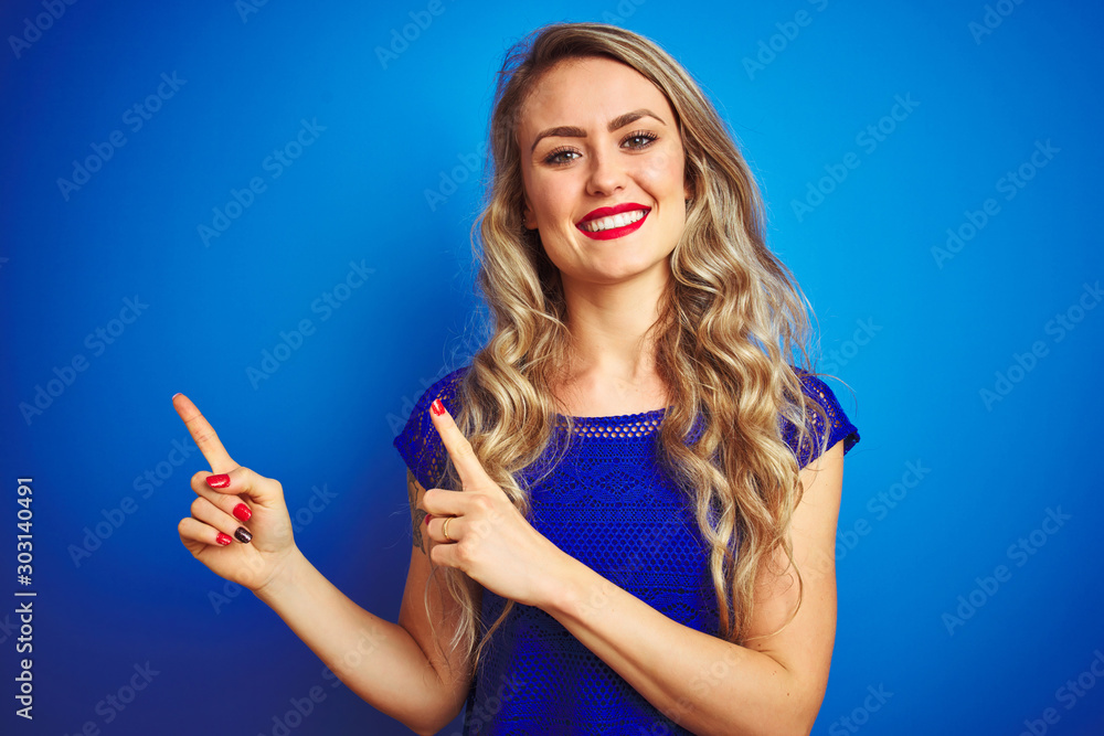 Obraz premium Young beautiful woman standing over blue isolated background smiling and looking at the camera pointing with two hands and fingers to the side.