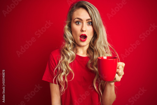 Young beautiful woman drinking a cup of cooffe over red isolated background scared in shock with a surprise face, afraid and excited with fear expression photo