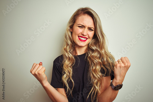 Young beautiful woman wearing black t-shirt standing over white isolated background very happy and excited doing winner gesture with arms raised, smiling and screaming for success. Celebration concept