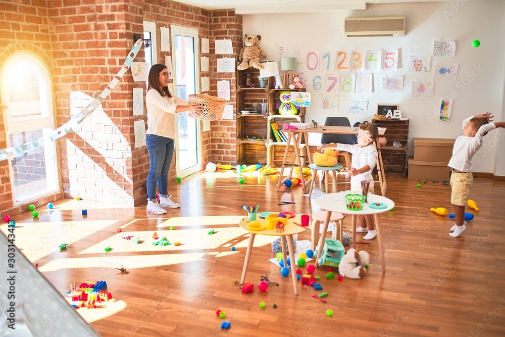 Obraz premium Beautiful teacher and toddlers playing basketball using balls and wicker basket around lots of toys at kindergarten