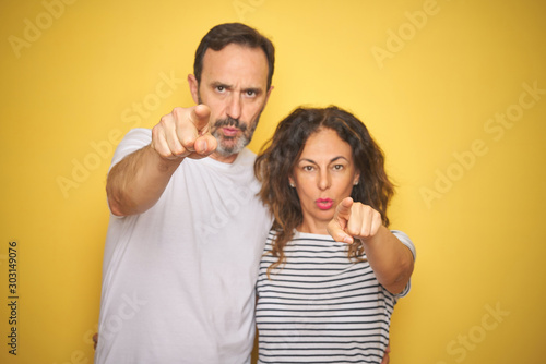 Beautiful middle age couple together standing over isolated yellow background pointing with finger to the camera and to you, hand sign, positive and confident gesture from the front