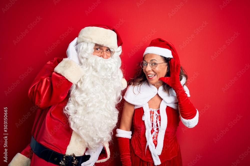 Middle age couple wearing Santa costume and glasses over isolated red background smiling with hand over ear listening an hearing to rumor or gossip. Deafness concept. Middle age couple wearing Santa costume and glasses over isolated red background smiling with hand over ear listening an hearing to rumor or gossip. Deafness concept.
