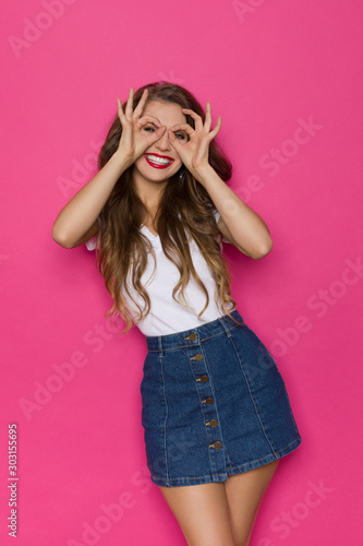 Beautiful Young Woman In Jeans Mini Skirt And White Top Is Making Hand Binoculars And Smiling. © studioloco