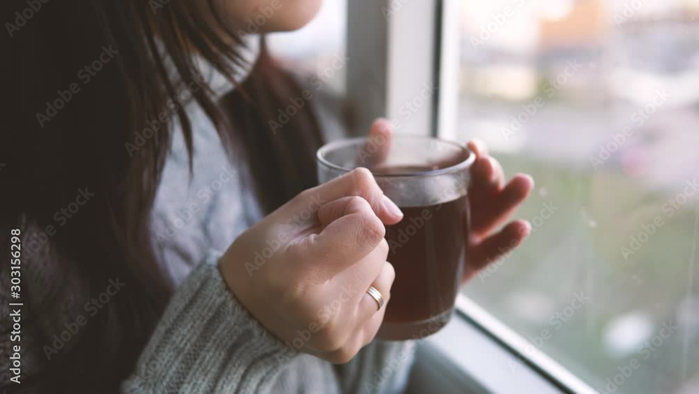 Close up, woman in a gray sweater drinks a tea, looking out the window on the balcony. Close up, woman in a gray sweater drinks a tea, looking out the window on the balcony.