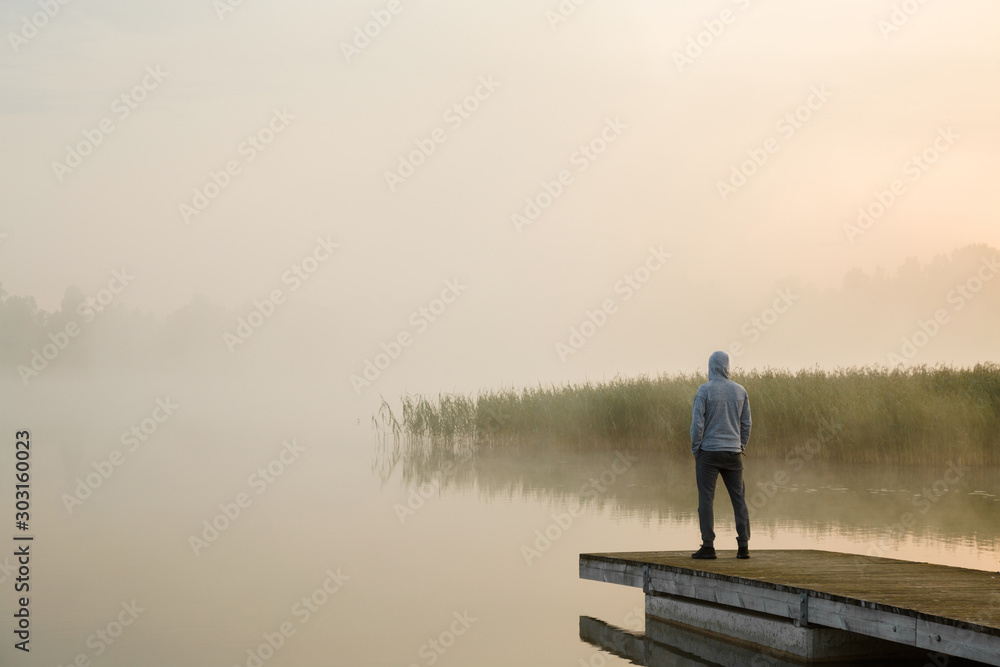 Young man standing alone on wooden footbridge and staring at lake. Mist over water. Foggy air. Early chilly morning. Empty place for sentimental, inspirational text, quote or sayings. Back view.