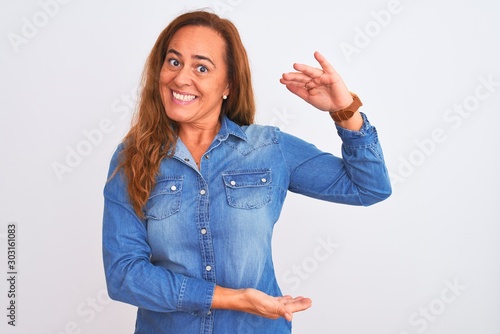 Middle age mature woman wearing denim jacket standing over white isolated background gesturing with hands showing big and large size sign, measure symbol. Smiling looking at the camera. Measuring