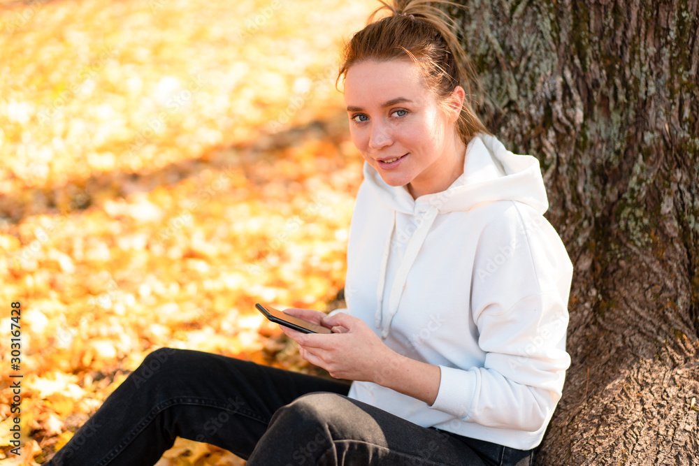 Obraz premium Woman in a white sweater with a hood sits on ground in the park near the tree and holding a cellphone in her hands