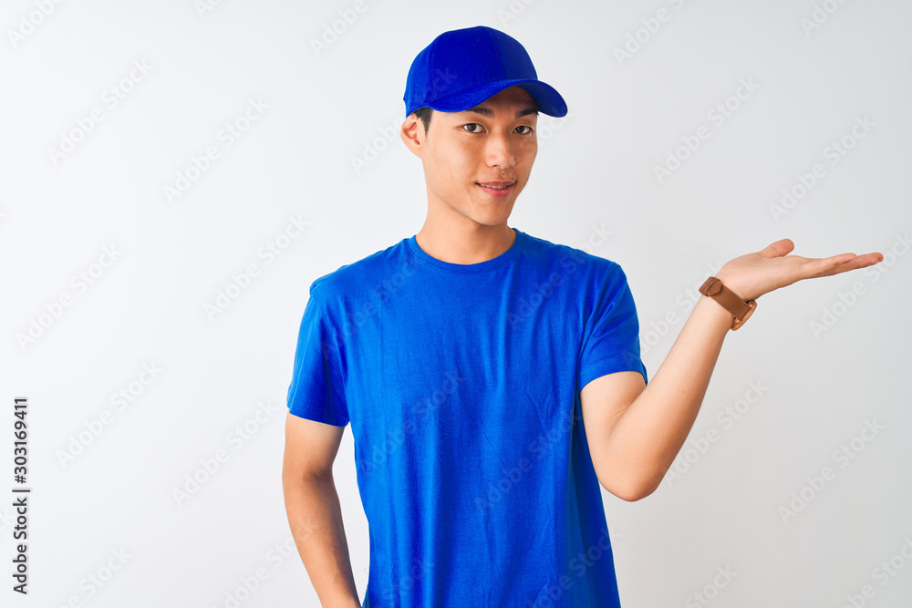 Chinese deliveryman wearing blue t-shirt and cap standing over isolated white background smiling cheerful presenting and pointing with palm of hand looking at the camera.