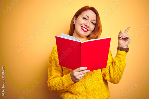 Young redhead teacher woman reading red book over yellow isolated background very happy pointing with hand and finger to the side