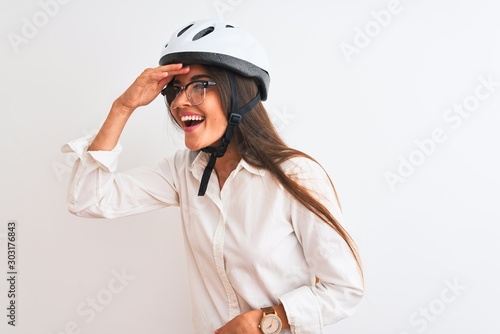 Beautiful businesswoman wearing glasses and bike helmet over isolated white background very happy and smiling looking far away with hand over head. Searching concept.