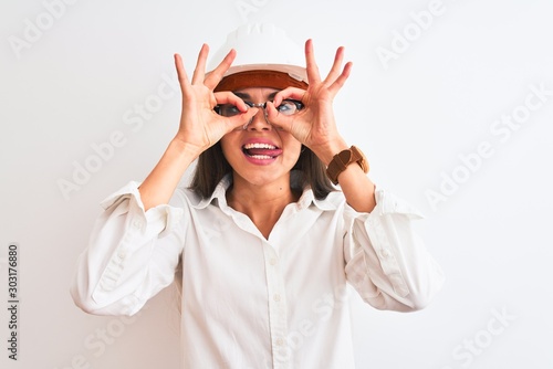 Young beautiful architect woman wearing helmet and glasses over isolated white background doing ok gesture like binoculars sticking tongue out, eyes looking through fingers. Crazy expression.