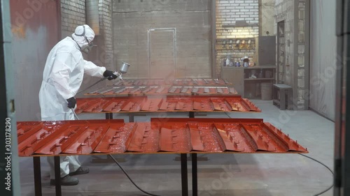 Professional worker at paint workshop works, using spray gun, applies coating composition to metal workpieces, red paint, spraying, painting, industrial production, spray from gun, spacious workshop