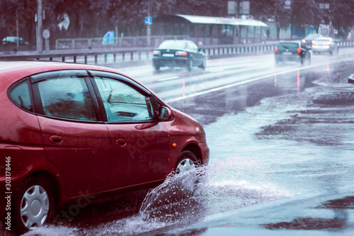 Ukraine. Kiev - 05,12,2019 Spraying water from the wheels of a vehicle moving on a wet city asphalt road. The wet wheel of a car moves at a speed along a puddle on a flooded city road during rain. © Yuliia