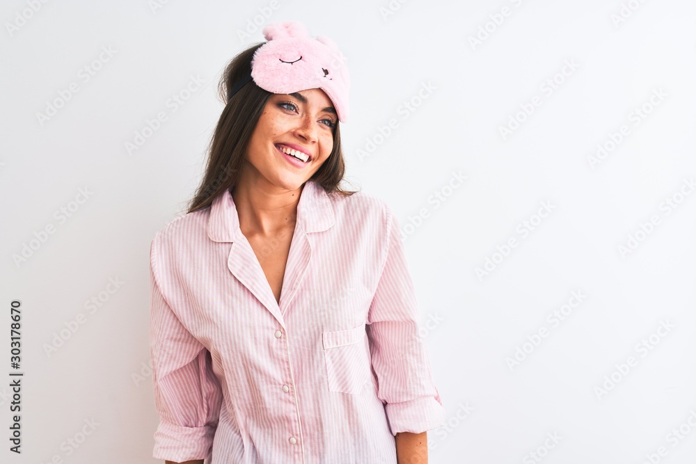 Young beautiful woman wearing sleep mask and pajama over isolated white background looking away to side with smile on face, natural expression. Laughing confident. Young beautiful woman wearing sleep mask and pajama over isolated white background looking away to side with smile on face, natural expression. Laughing confident.