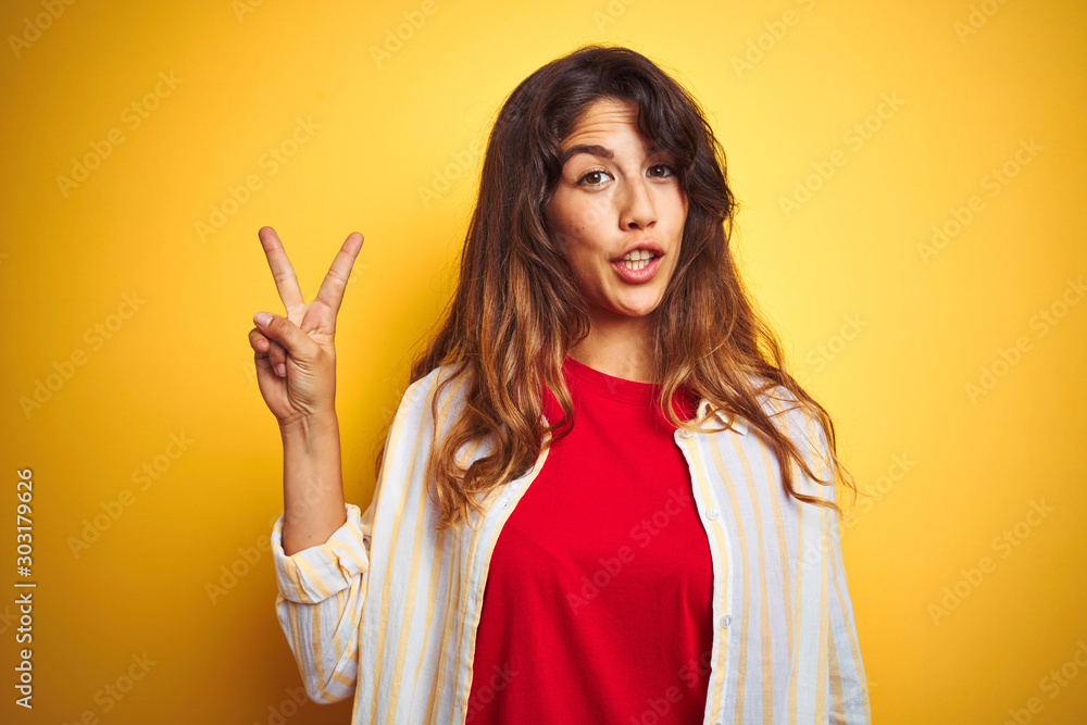 Young beautiful woman wearing red t-shirt and stripes shirt over yellow isolated background showing and pointing up with fingers number two while smiling confident and happy. Young beautiful woman wearing red t-shirt and stripes shirt over yellow isolated background showing and pointing up with fingers number two while smiling confident and happy.
