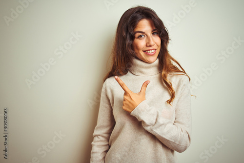 Young beautiful woman wearing winter sweater standing over white isolated background cheerful with a smile on face pointing with hand and finger up to the side with happy and natural expression © Krakenimages.com