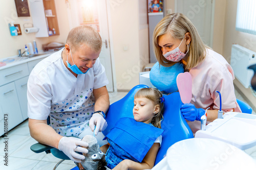 Dentist examining little girl's teeth in the stomatology chair with assistant's help. Doctor holds a soft toy. Selective focus. Pediatric stomatology concept.