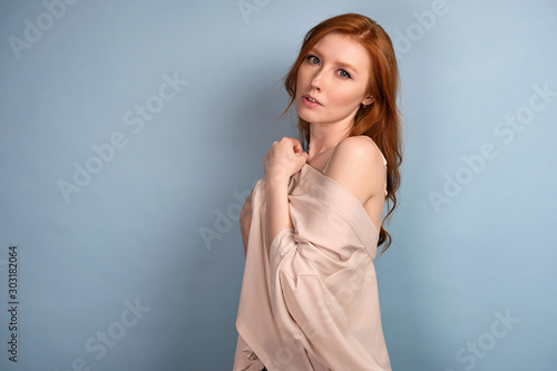 Red-haired girl stands in profile on a blue background in a light robe lowered from the shoulders.