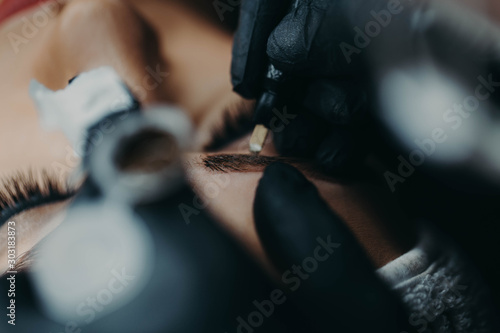 Microblading master in black rubber gloves with a needle on the manipulator applies pigment to the eyebrows of the model close-up.