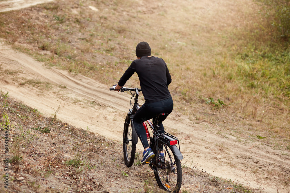 Outdoor shot of male bike cyclist riding uphill along country road, spending clear day in active way, sporty guy covering assigned distance, riding uphill, man wearing black track suit, back view.