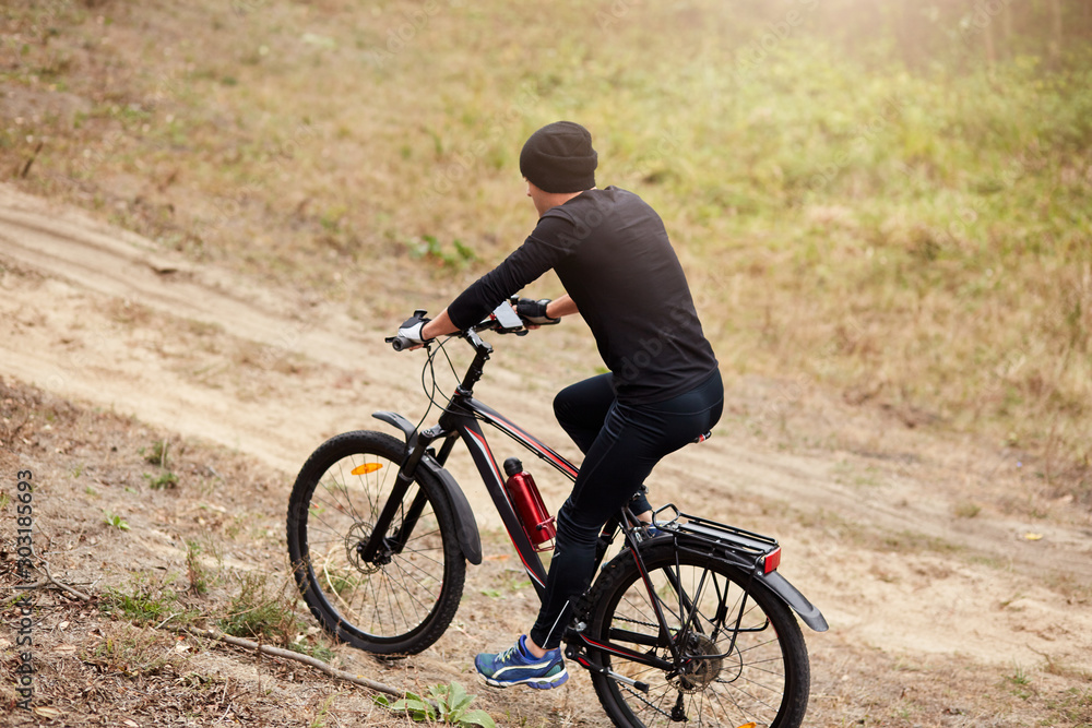 Side wayof male mountain bike cyclist riding uphill along country road, guy wearing black sportwear and cap, sportsman covering assigned distance, enjoying his hobby during vacation. Sport concept. Side wayof male mountain bike cyclist riding uphill along country road, guy wearing black sportwear and cap, sportsman covering assigned distance, enjoying his hobby during vacation. Sport concept.