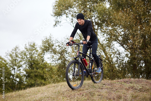 Attaractive man wearing blacktrack suit and cap riding dowhill on his mountain bike  having cardio training  enjoying his recreating in open air and beautiful nature. Sport  healthy lifestyle concept.
