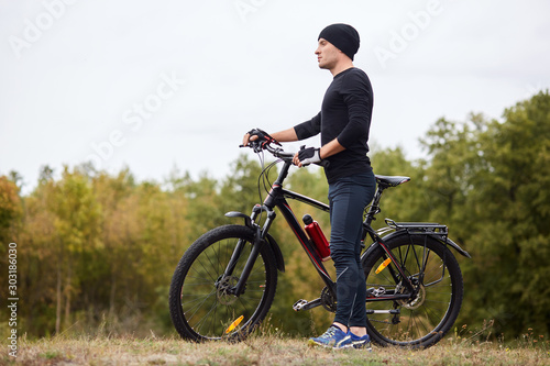Active sportsman cyclist dresses black track suit standing with bikes on hill near green trees, enjoying view in front of him, malebeing good at moutain biking and road cycling. Sport concept.