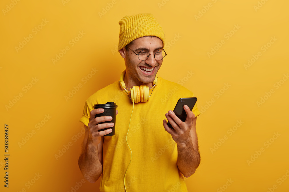 Cheerful smiling guy looks funny video via smartphone, drinks tasty hot beverage from paper cup, wears yellow hat and t shirt, uses headphones for listening music. People and technology concept Cheerful smiling guy looks funny video via smartphone, drinks tasty hot beverage from paper cup, wears yellow hat and t shirt, uses headphones for listening music. People and technology concept
