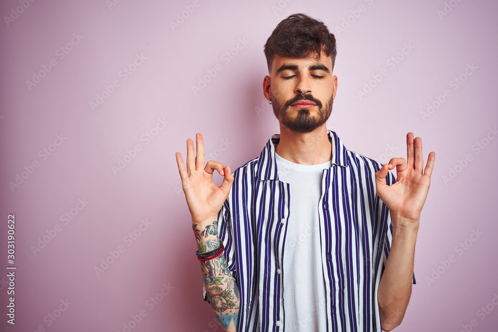 Naklejka premium Young man with tattoo wearing striped shirt standing over isolated pink background relax and smiling with eyes closed doing meditation gesture with fingers. Yoga concept.