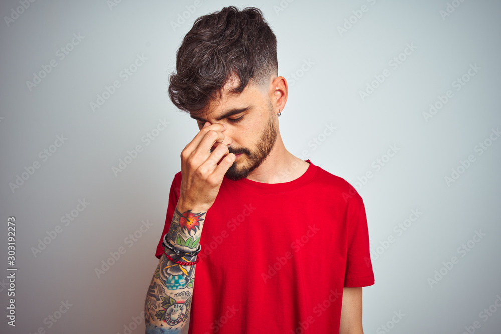 Young man with tattoo wearing red t-shirt standing over isolated white background tired rubbing nose and eyes feeling fatigue and headache. Stress and frustration concept. Young man with tattoo wearing red t-shirt standing over isolated white background tired rubbing nose and eyes feeling fatigue and headache. Stress and frustration concept.