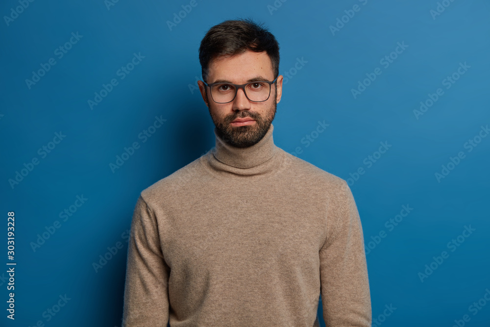Portrait of serious handsome male has dark hair, thick bristle, looks straightly at camera, wears optical glasses and turtleneck jumper, isolated over blue background, listens attentively interlocutor Portrait of serious handsome male has dark hair, thick bristle, looks straightly at camera, wears optical glasses and turtleneck jumper, isolated over blue background, listens attentively interlocutor