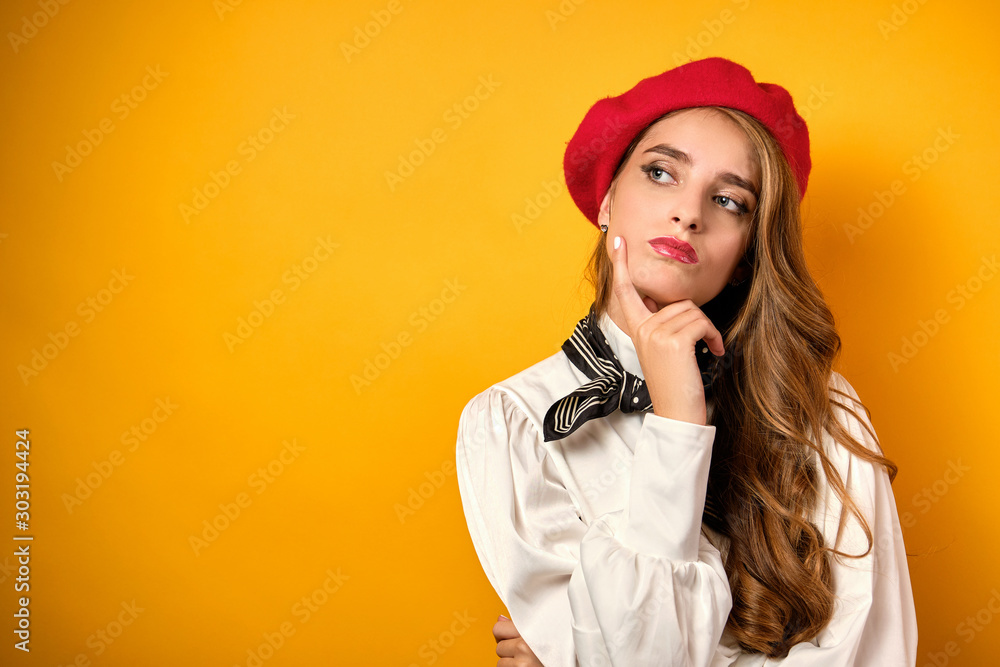A beautiful blonde with red lips in a white blouse and a red beret looks thoughtfully at the side, pressing a finger to her face. A beautiful blonde with red lips in a white blouse and a red beret looks thoughtfully at the side, pressing a finger to her face.