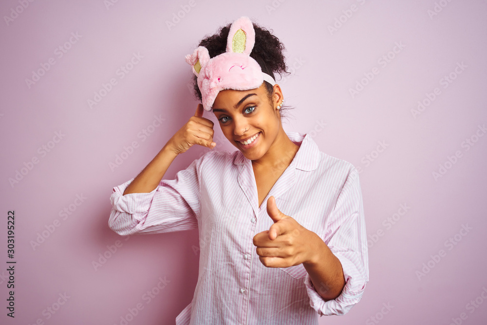 Young african american woman wearing pajama and mask over isolated pink background smiling doing talking on the telephone gesture and pointing to you. Call me.