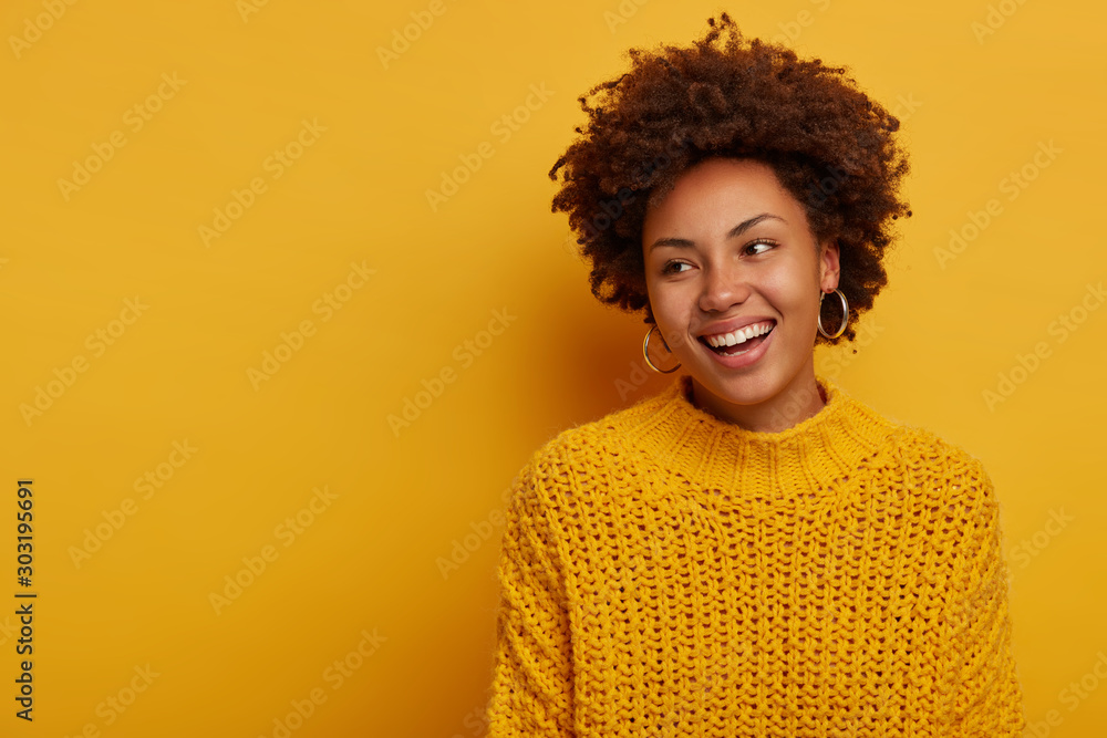 Tender charming happy curly woman has relaxed joyful face expression, Afro hairstyle, wears knitted sweater, laughs enthusiastic, poses against yellow background, blank space aside, turns away - obrazy, fototapety, plakaty 