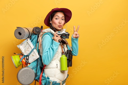 Tableau sur toile Pretty active backpacker makes victory gesture, keeps lips rounded, holds retro camera, stands with travelbag, takes photos during trip, isolated over yellow background