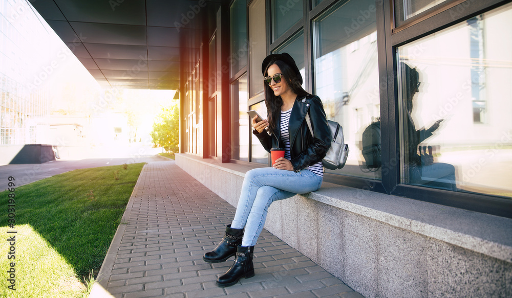 Like a boss. Full-length photo of charming lady in a fashionable clothes, who is sitting near a modern building with a smartphone, drinking her coffee on the go. Like a boss. Full-length photo of charming lady in a fashionable clothes, who is sitting near a modern building with a smartphone, drinking her coffee on the go.