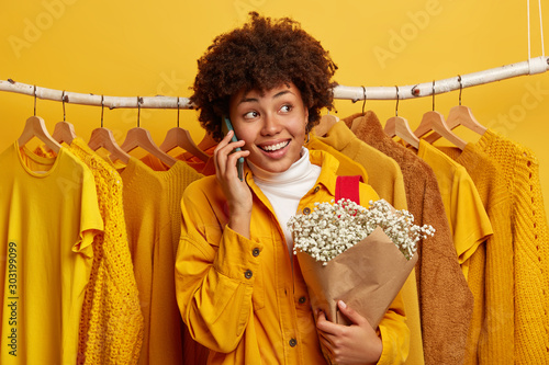 Photo of delighted curly woman turns away, calls via cellular, shares impressions after shopping day, poses with bouquet of flowers, poses against bright clothes on hangers, isolated on yellow wall photo