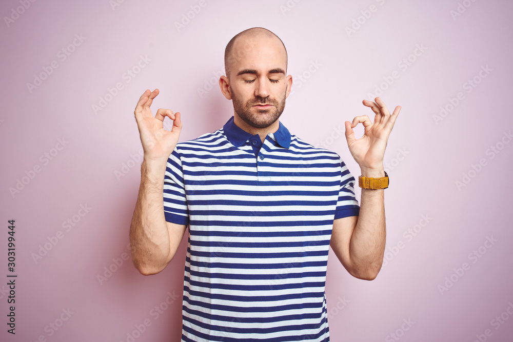 Fototapeta premium Young bald man with beard wearing casual striped blue t-shirt over pink isolated background relax and smiling with eyes closed doing meditation gesture with fingers. Yoga concept.