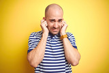Young bald man with beard wearing casual striped blue t-shirt over yellow isolated background covering ears with fingers with annoyed expression for the noise of loud music. Deaf concept.