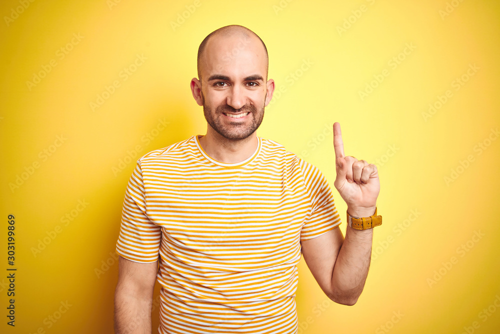 Young bald man with beard wearing casual striped t-shirt over yellow isolated background showing and pointing up with finger number one while smiling confident and happy.