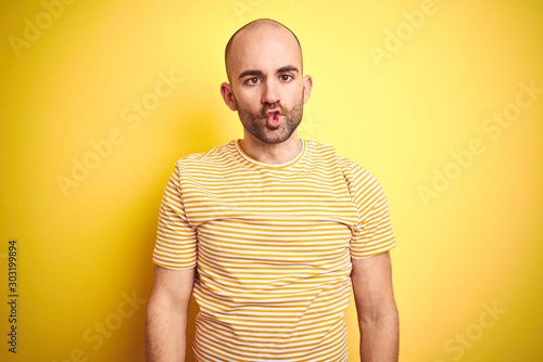 Young bald man with beard wearing casual striped t-shirt over yellow isolated background making fish face with lips, crazy and comical gesture. Funny expression.